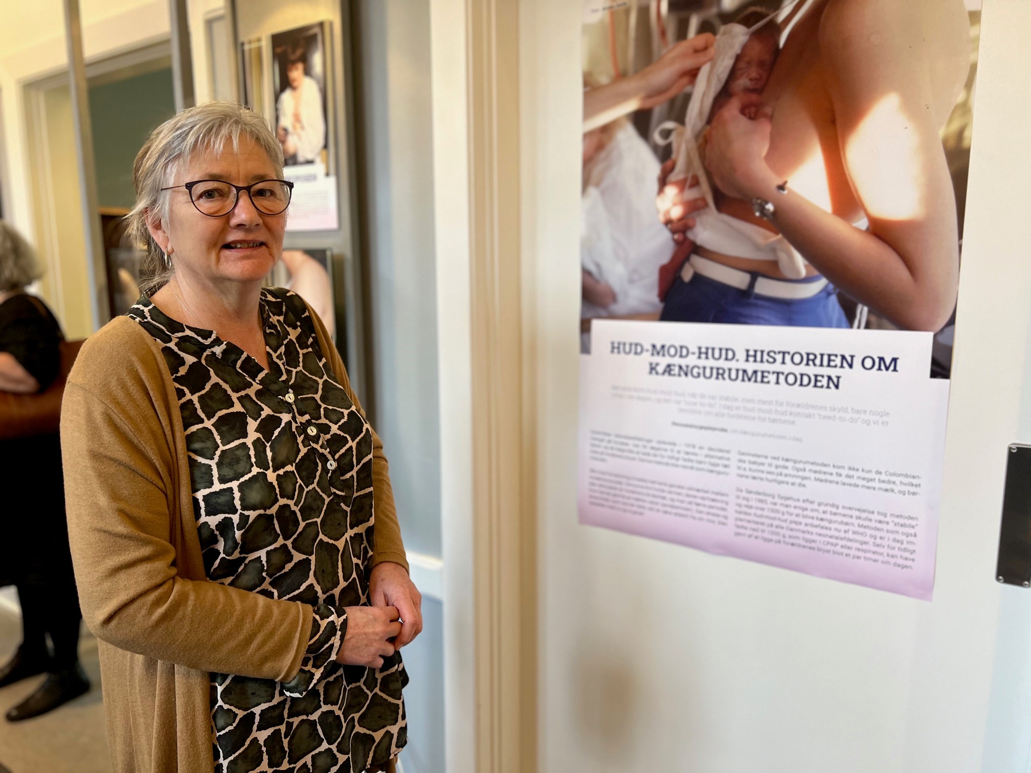 Sygeplejerske Kirsten Hjort-Gregersen foran en af plancherne på udstillingen. Kirsten blev ansat som sygeplejerske på Neonatal-afdelingen i Sønderborg i 1981. Hun husker tydeligt, da man i 1985 som noget nyt, valgte at mødre og børn fik mulighed for at ligge på samme afdeling.